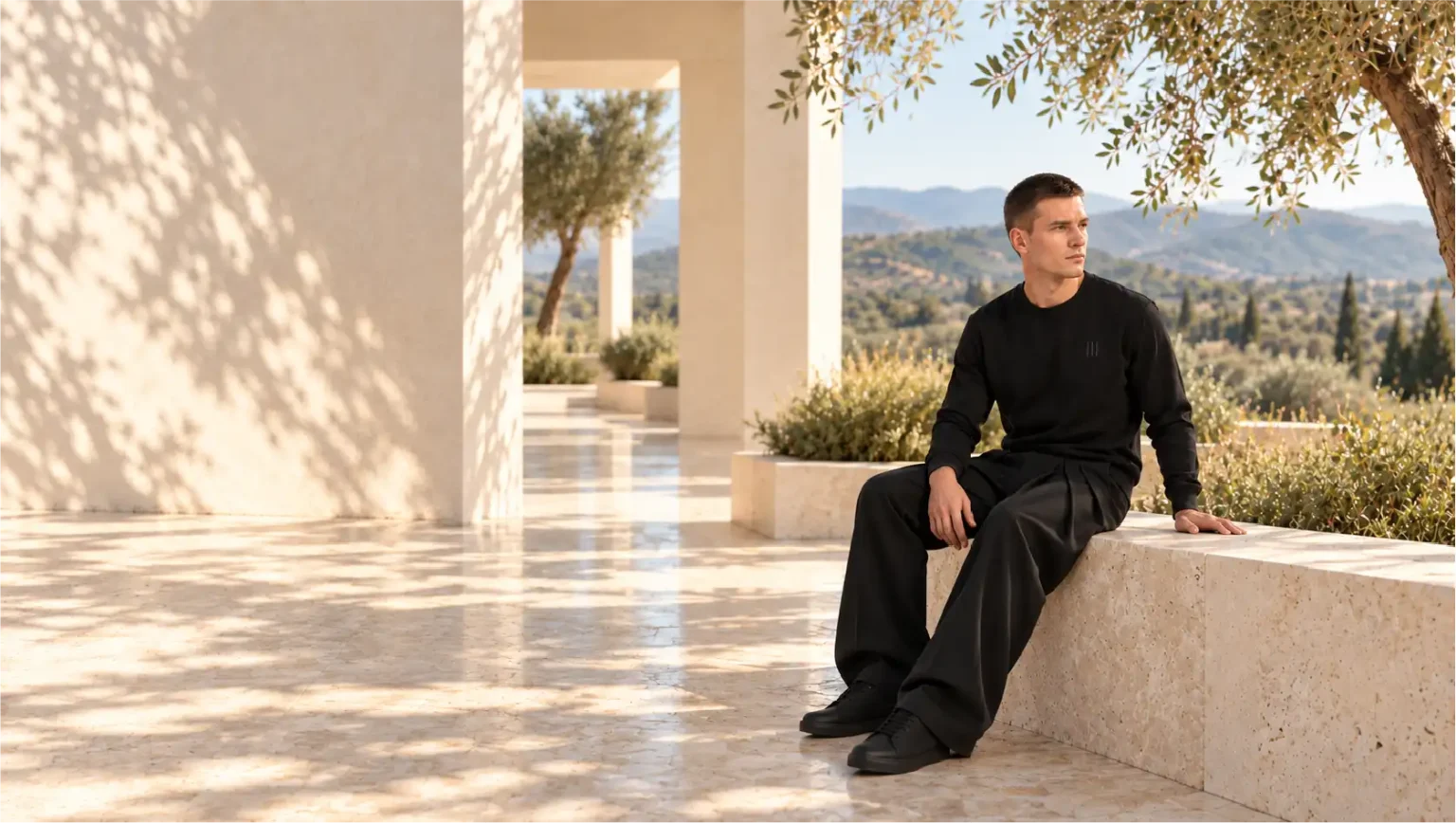 Model seated on a stone wall in a sunlit courtyard, wearing a black sweatshirt with fine vertical back artwork and loose black trousers.