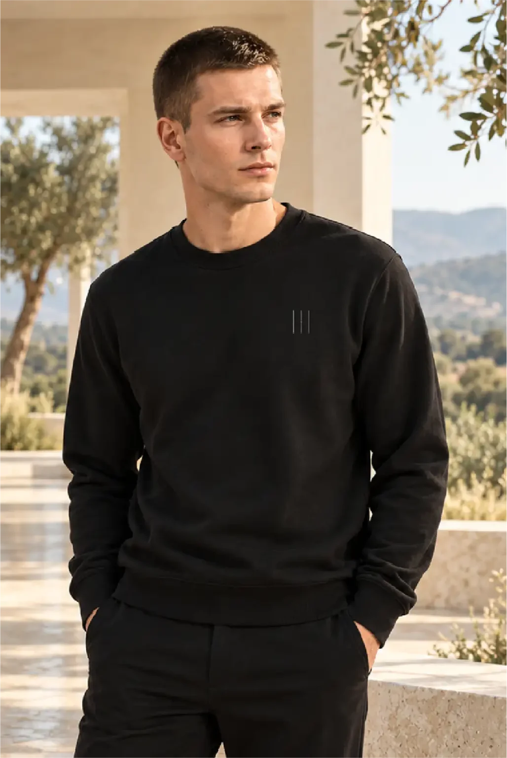 Model standing in a sunlit courtyard, wearing a black sweatshirt with fine vertical front artwork and loose black trousers.
